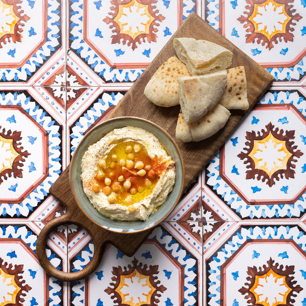 Bowl of hummus with pita bread on a wooden board against a colourful tiled table.